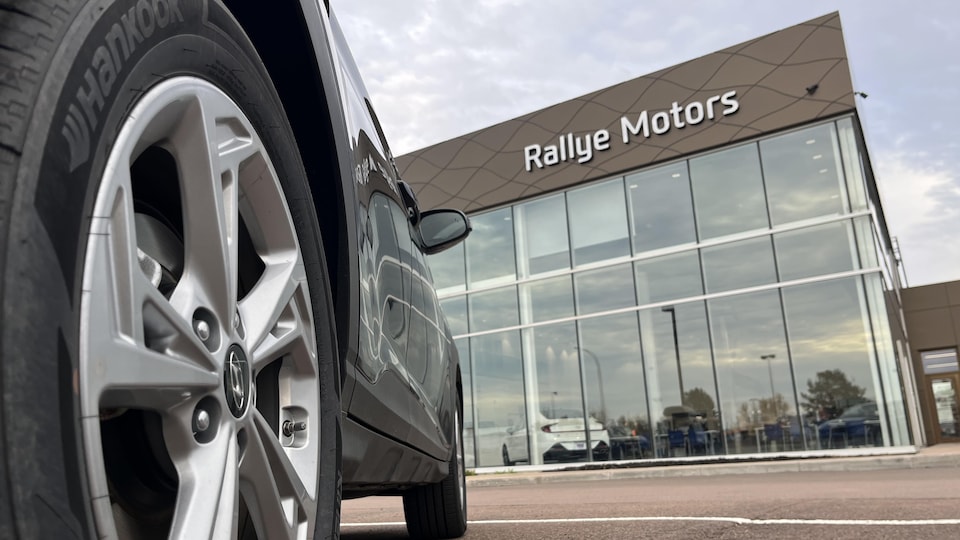 Une voiture stationnée devant un édifice de Rallye Motors. La photo est prise d’un angle où l’on voit le pneu arrière en gros plan. 
